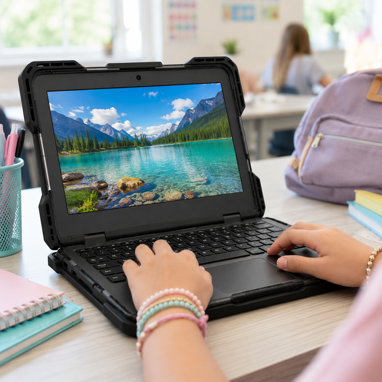InfoCase Rugged Pro protective laptop case shown in use on a light classroom desk, with a student wearing bracelets using the device and a scenic image displayed on screen.